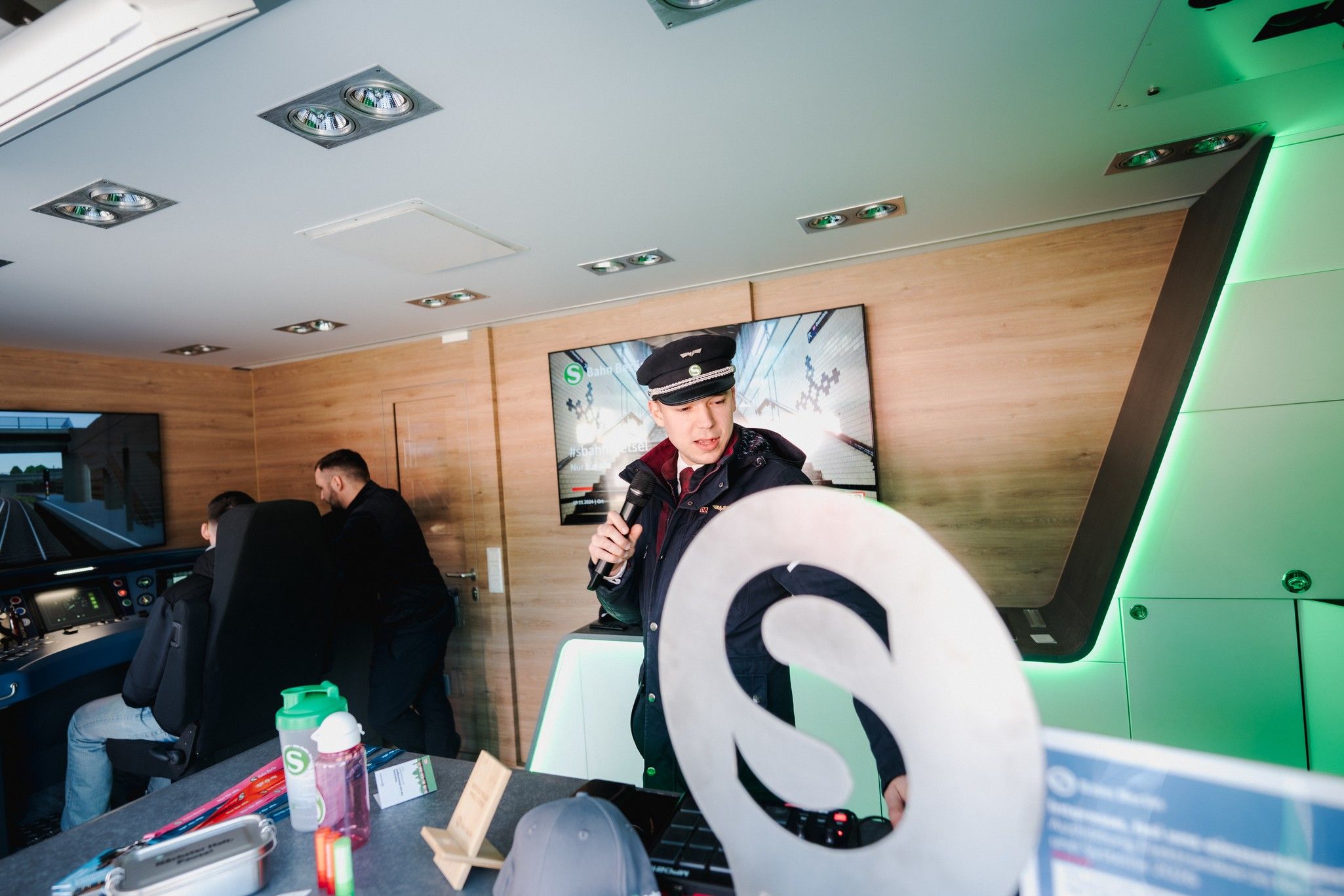 Eine Person mit Kappe spricht ins Mikrofon in einem modernen Zugwagen, während zwei weitere Personen im Hintergrund sitzen.