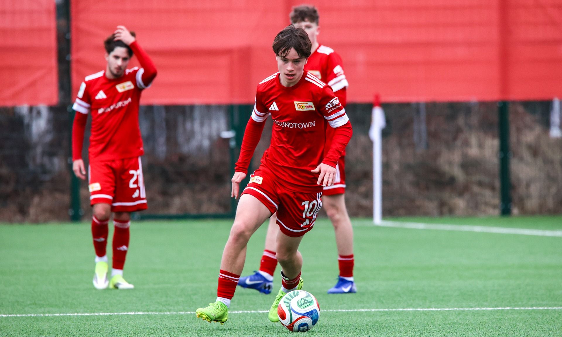 Ein junger Fußballspieler in rotem Trikot dribbelt den Ball auf einem Trainingsplatz, während andere Spieler im Hintergrund stehen.