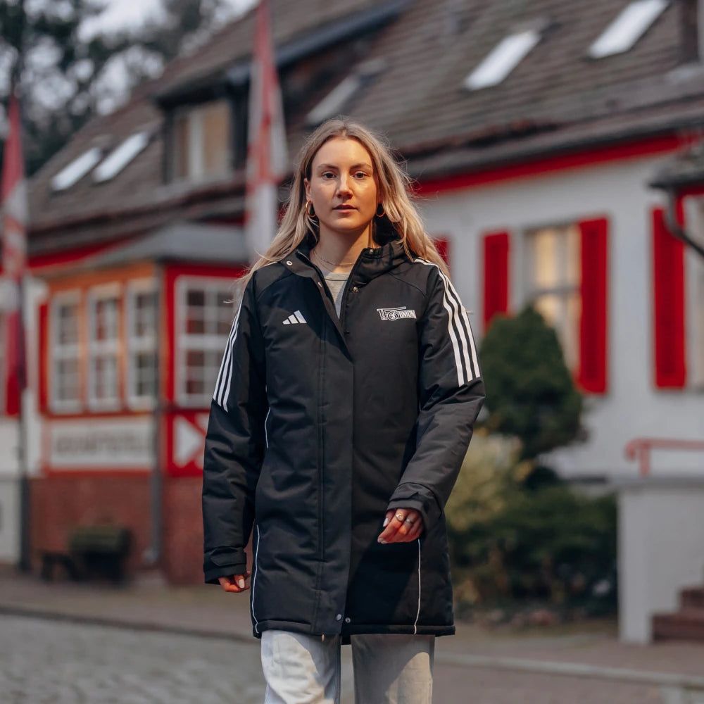 Junge Frau in schwarzer Sportjacke mit weißen Streifen, läuft auf einer Straße vor einem Gebäude mit roten Fenstern.