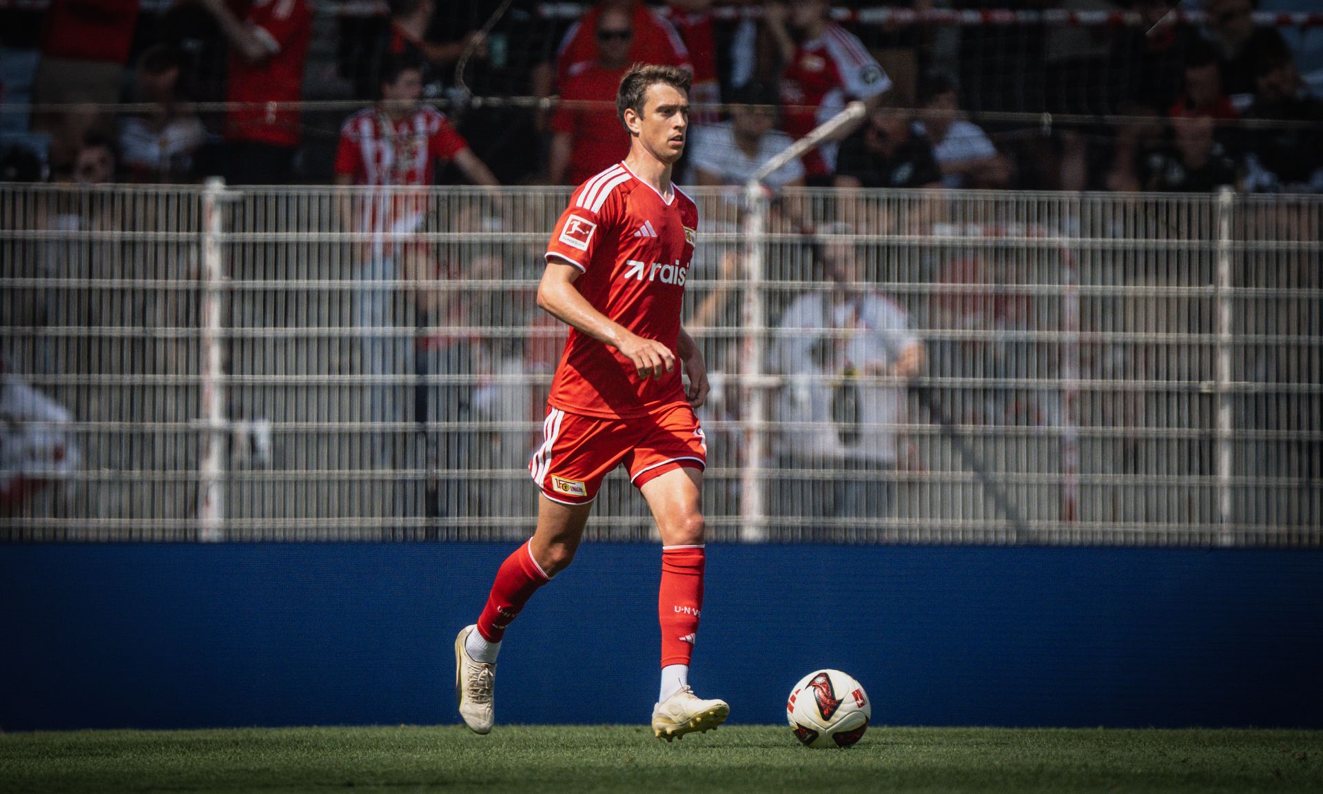 Spieler in rotem Trikot läuft mit einem Fußball auf dem Platz, im Hintergrund Zuschauer im Stadion.