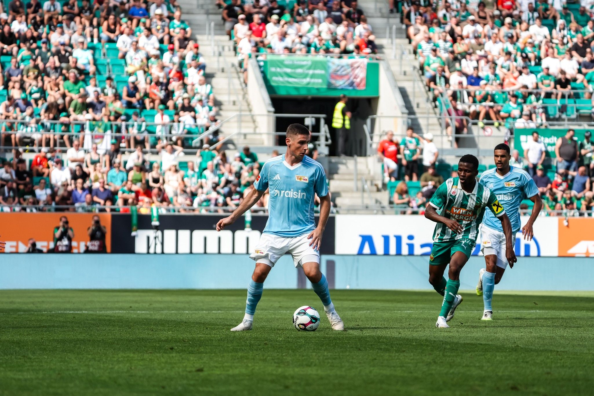 Fußballspiel mit Spielern in blauen und grünen Trikots, umgeben von einer Menschenmenge auf den Tribünen.