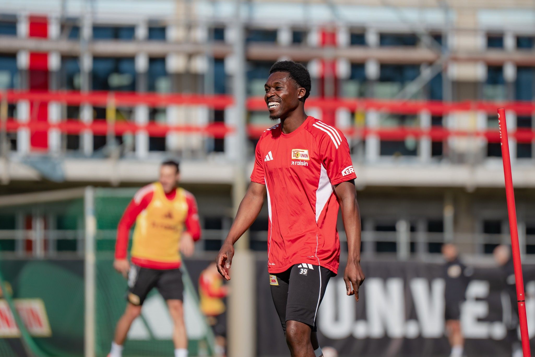 Ein Fußballspieler in rotem Trikot lächelt beim Training, während im Hintergrund weitere Spieler aktiv sind.