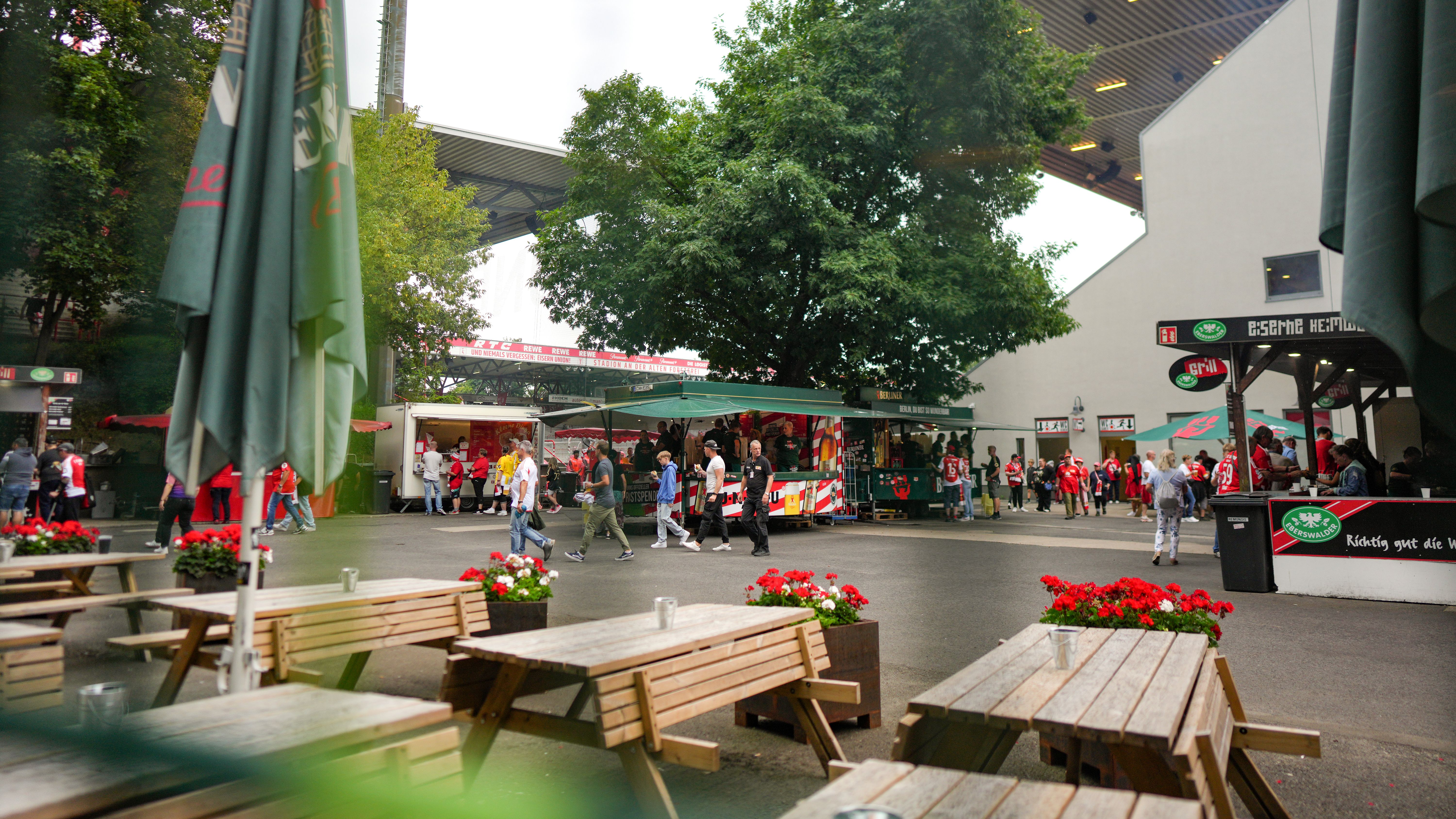 Belebter Außenbereich eines Stadions mit Essensständen, Tischen und Gästen in Sportbekleidung. Grüne Bäume im Hintergrund.