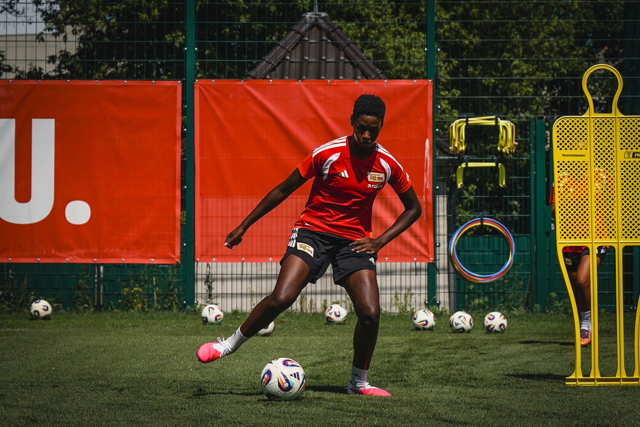Eine Person in einem roten Fußballtrikot trainiert auf einem Rasenplatz, umgeben von Fußballen und Trainingsgeräten.