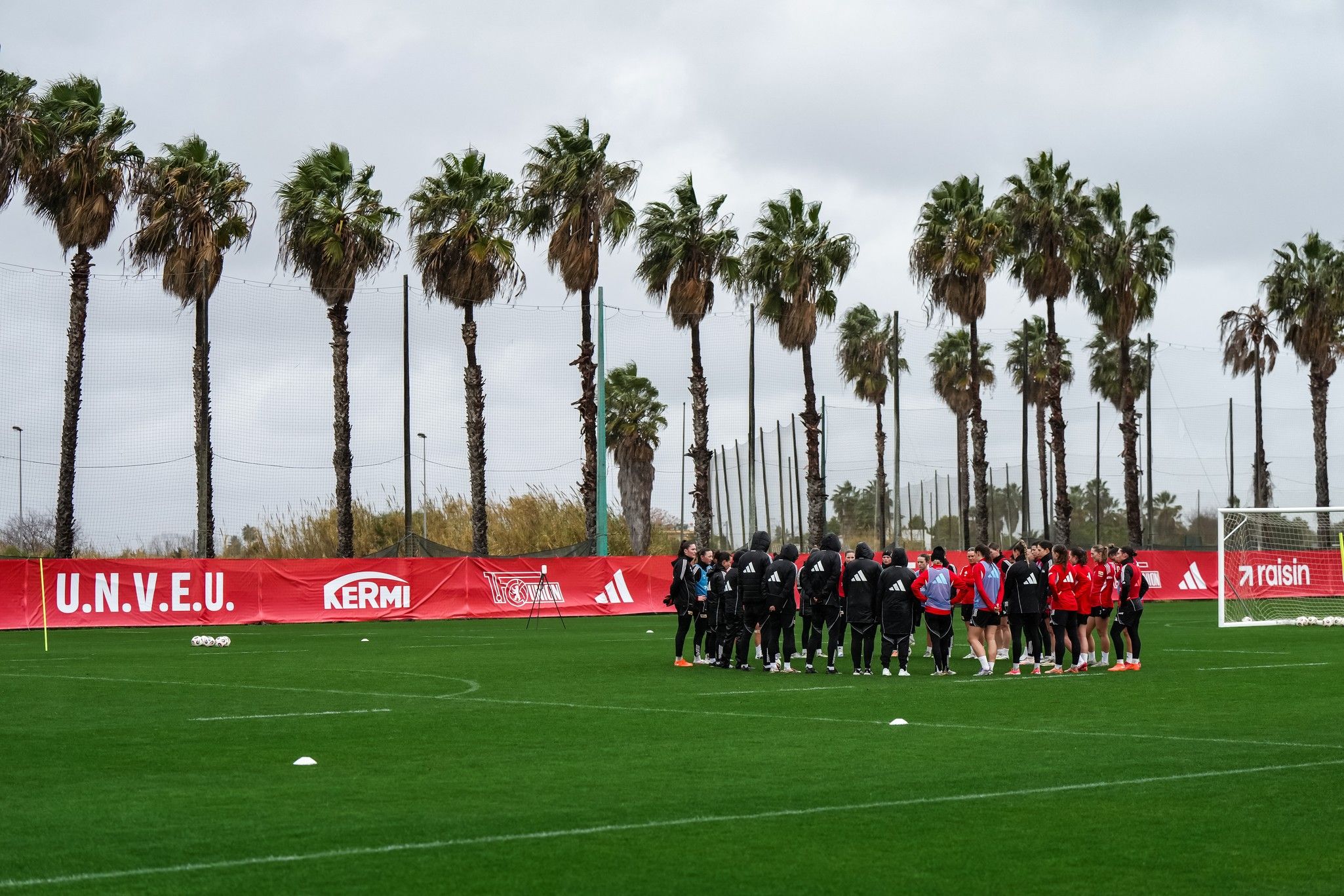 Gruppe von Personen auf einem Fußballplatz, umgeben von Palmen und einer Werbebande im Hintergrund.