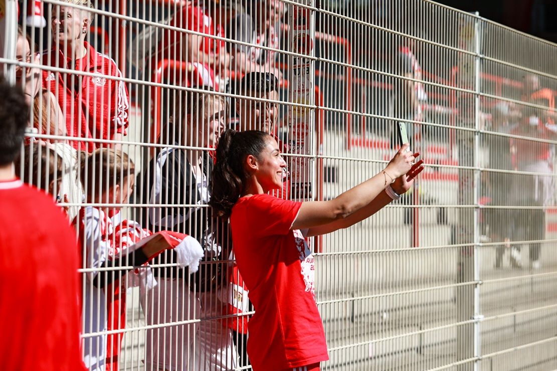 Eine Gruppe von Fans in roten Trikots macht Fotos hinter einem Zaun in einem Stadion.