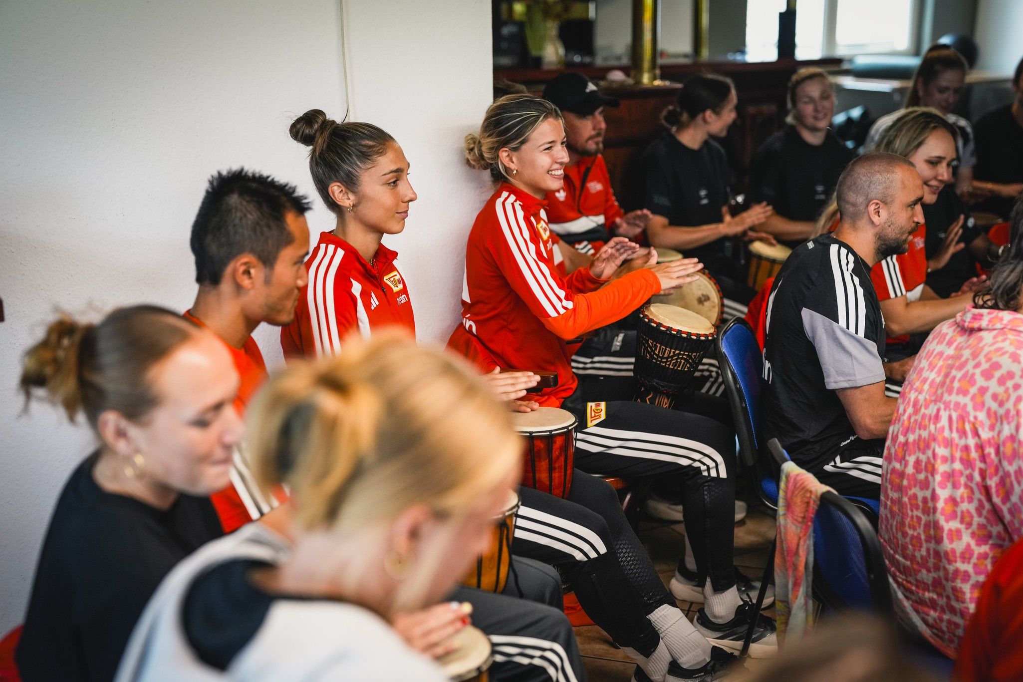Gruppensitzung mit mehreren Personen in sportlicher Kleidung, die auf Trommeln spielen und lächeln.