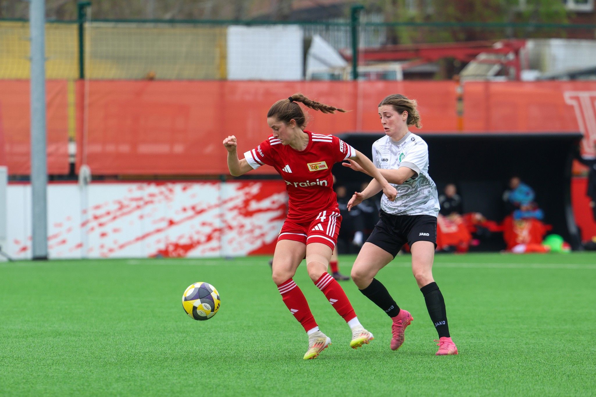 Zwei Spielerinnen in roten und weißen Trikots kämpfen um den Ball auf einem Fußballfeld.