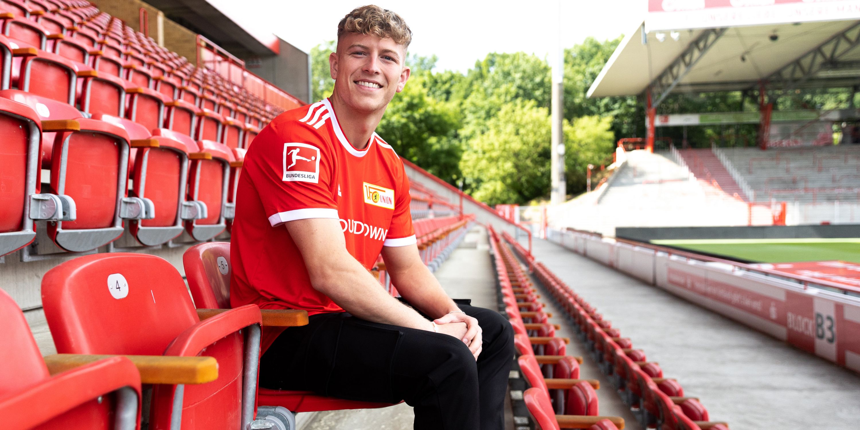 A young man in a red football jersey is sitting smiling in an empty stadium seat.