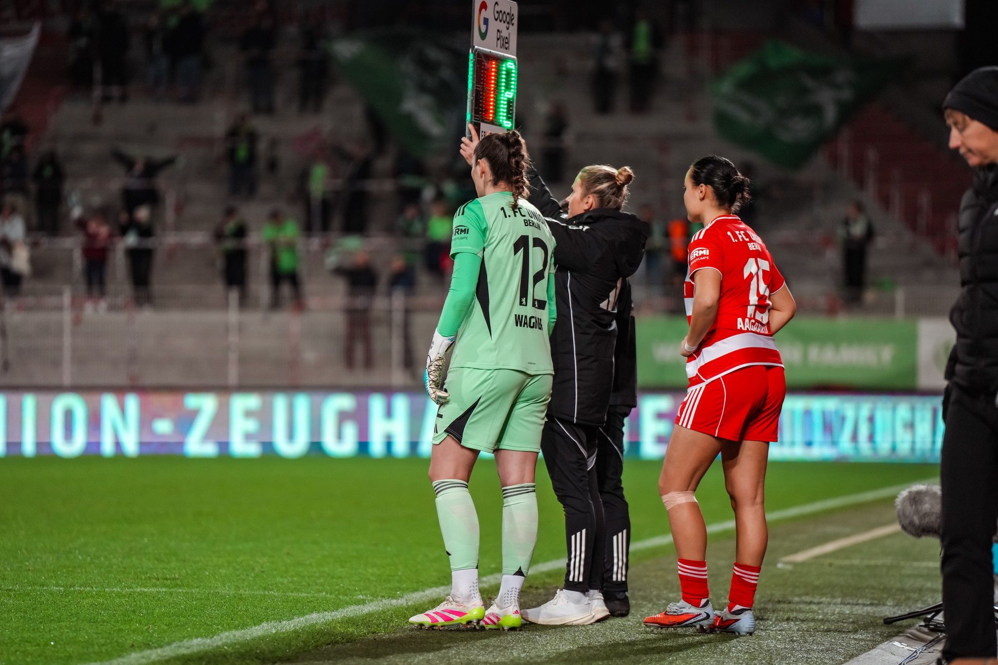 Torhüterin steht an der Seitenlinie, während eine Auswechslung im Fußballspiel vorbereitet wird. Zuschauer im Hintergrund sind zu sehen.