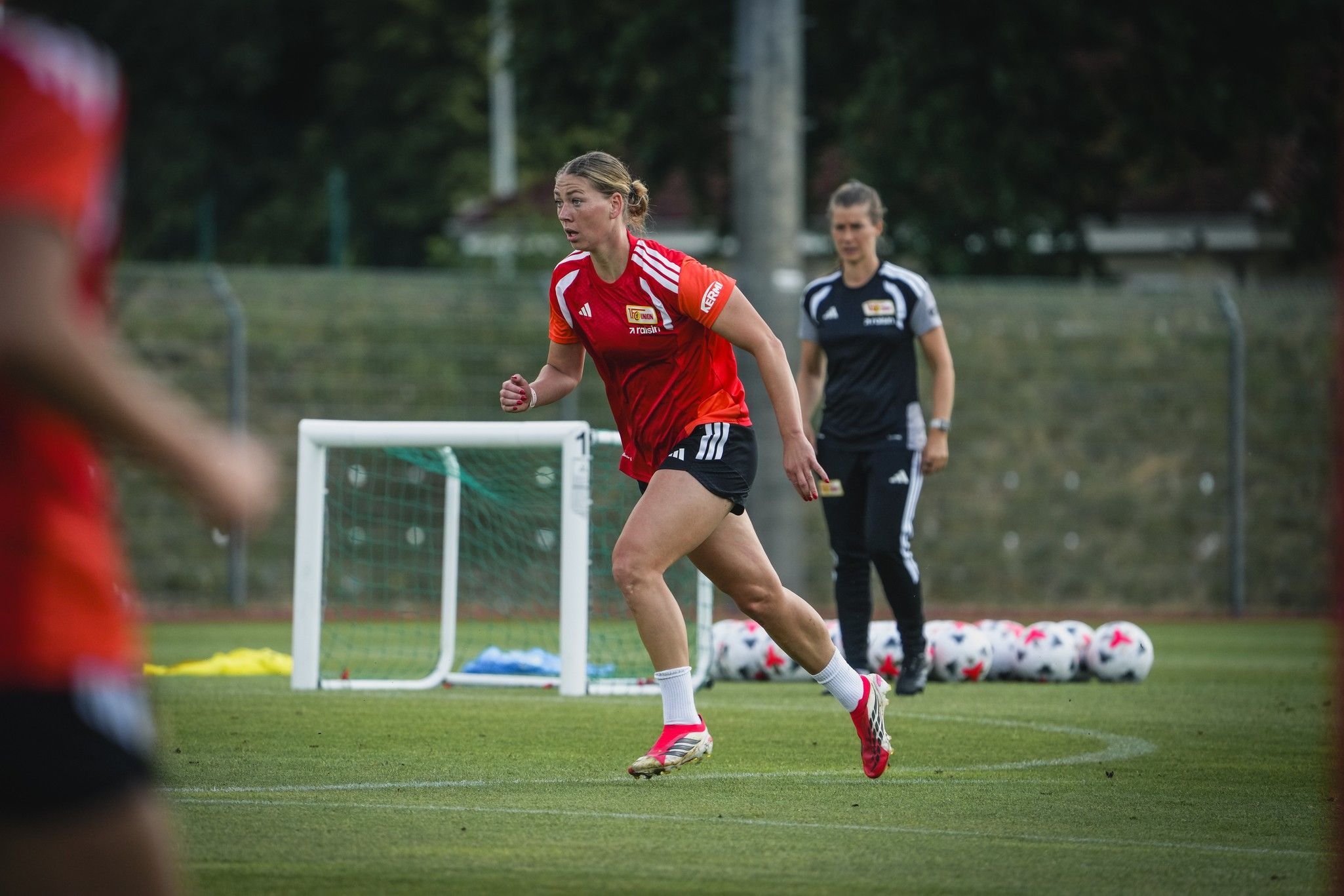 Eine Spielerin in rotem Trikot trainiert auf dem Fußballfeld, während eine Trainerin im Hintergrund zusieht.
