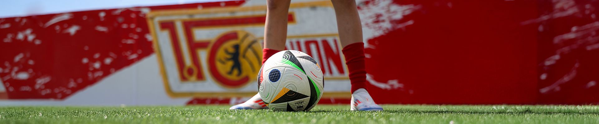 Ein Fußball liegt auf grünem Rasen, während die Füße eines Spielers mit roten Socken daneben stehen.