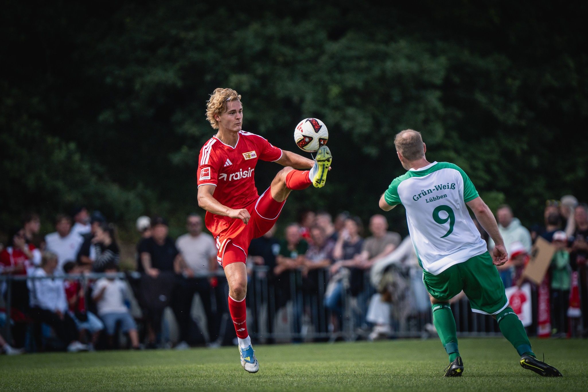 Ein Fußballspieler in rotem Trikot hebt das Bein, um den Ball zu spielen, während ein Gegner in grün-weiß zuschaut. Zuschauer im Hintergrund.