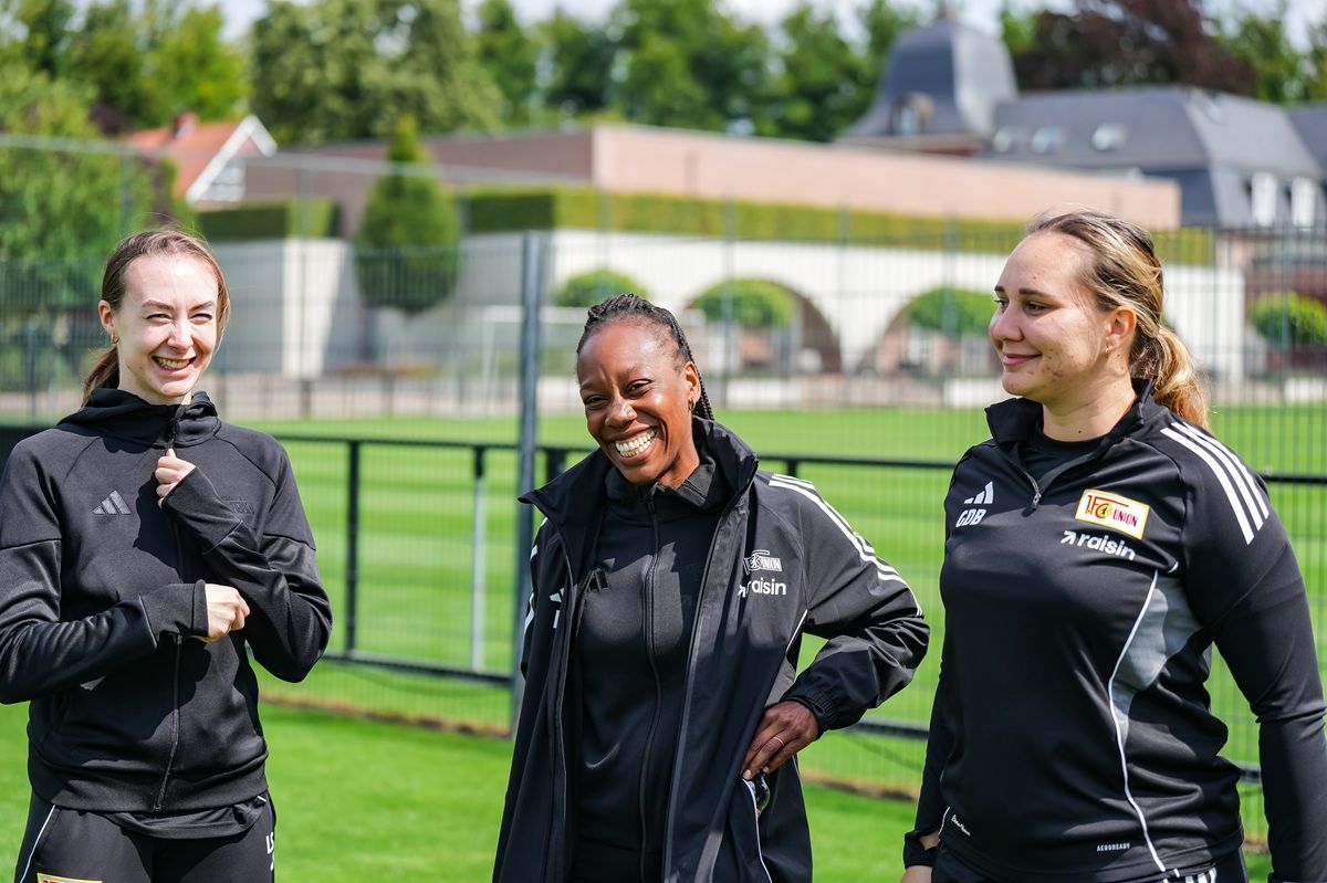 Drei Sportlerinnen in Trainingsbekleidung stehen lachend auf einem Fußballplatz und unterhalten sich.