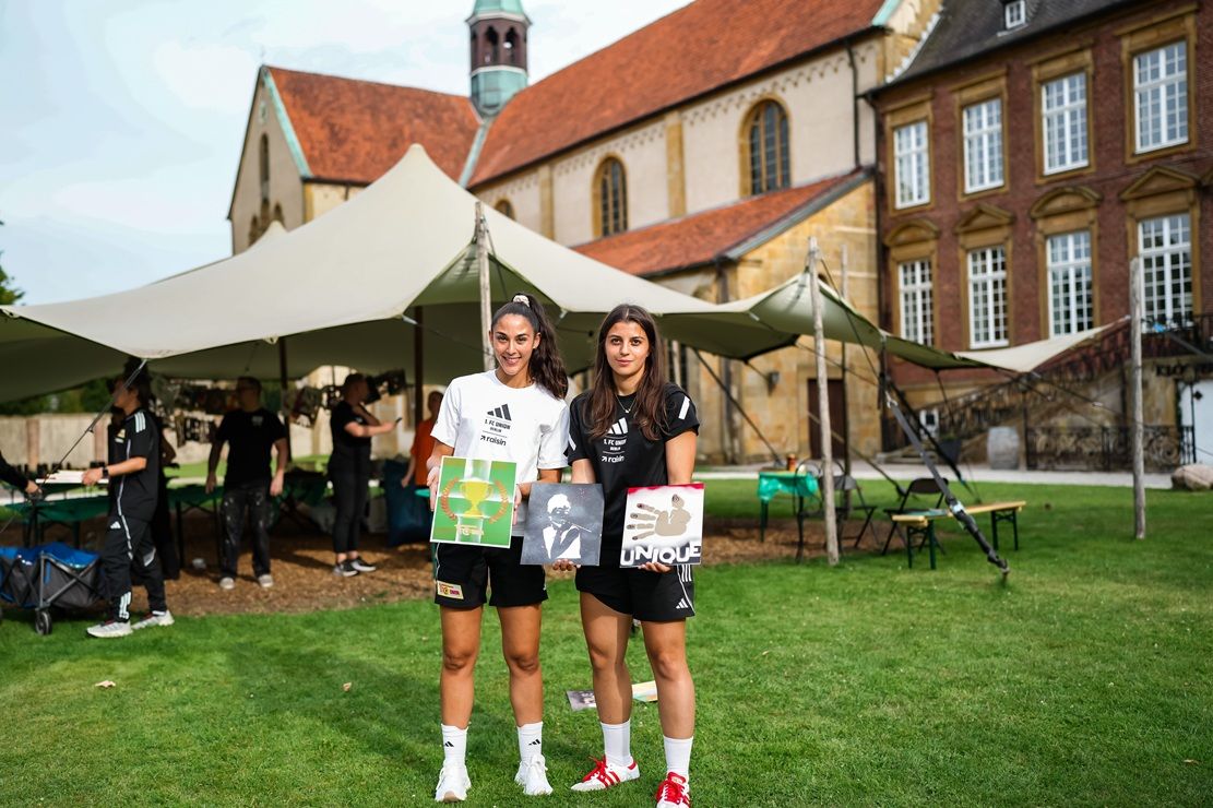 Zwei Fußballspielerinnen stehen im Freien und halten Kunstwerke, mit einem Zelt und historischen Gebäuden im Hintergrund.