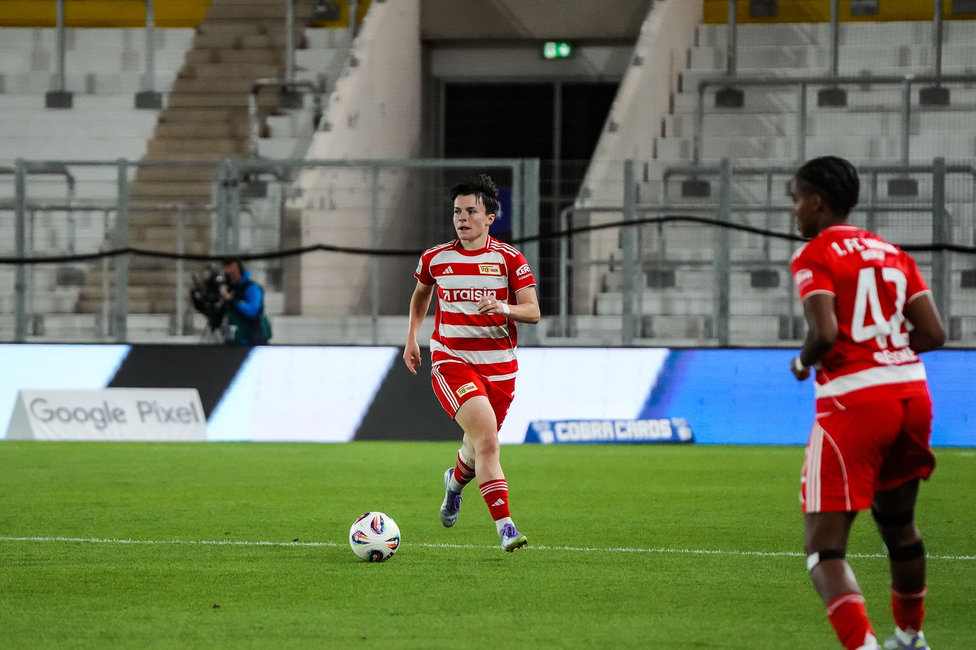 Spielszene im Fußball mit einer Spielerin in rotem Trikot, die den Ball führt, während eine Mitspielerin sie unterstützt.