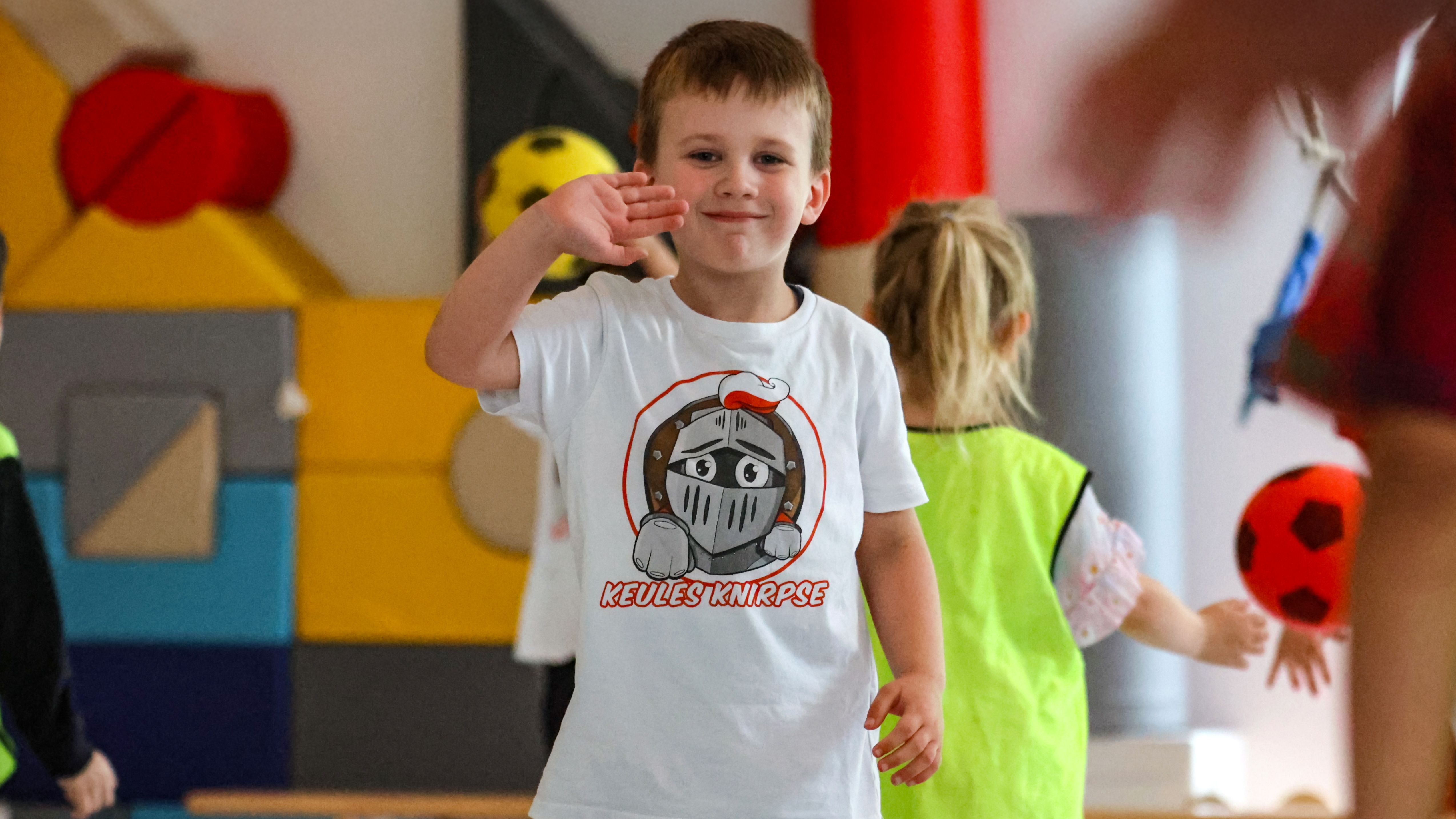 Ein Junge in einem weißen T-Shirt mit einem grafischen Aufdruck winkt fröhlich, umgeben von anderen Kindern in einer Spielumgebung.