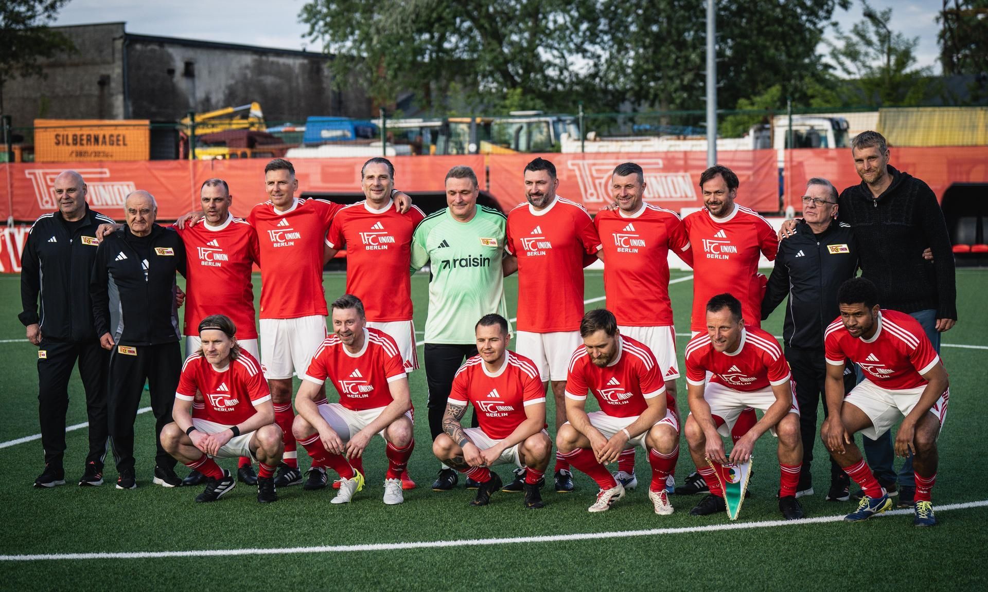 Gruppenfoto einer Fußballmannschaft in roten Trikots auf einem Sportplatz, umgeben von Trainern und Betreuern.