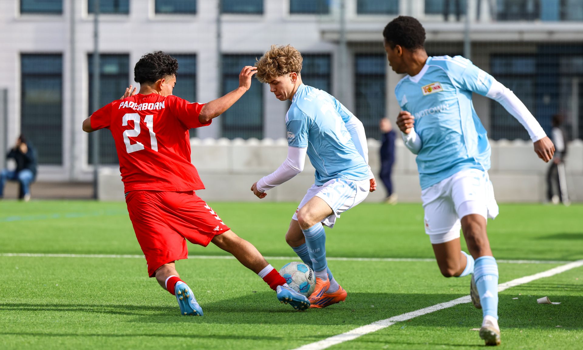 Zwei Spieler in roten und blauen Trikots kämpfen um den Ball auf einem Fußballplatz.