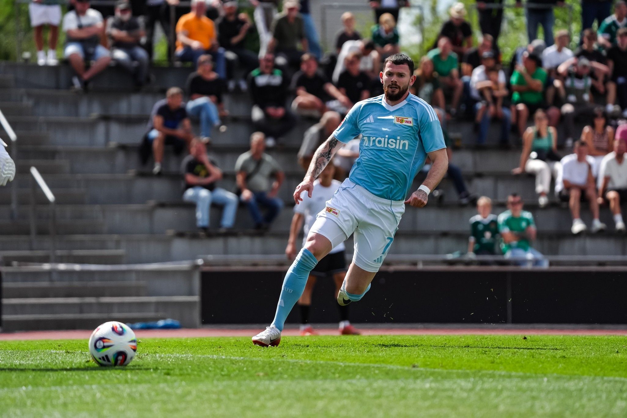 Ein Fußballspieler in hellblauem Trikot läuft auf das Spielfeld, während Zuschauer auf Tribünen zuschauen.