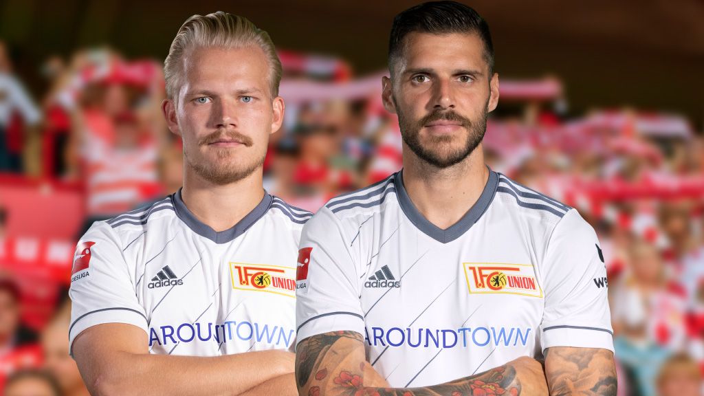 Zwei Fußballspieler im weißen Trikot des 1. FC Union Berlin stehen vor einer jubelnden Zuschauermenge.
