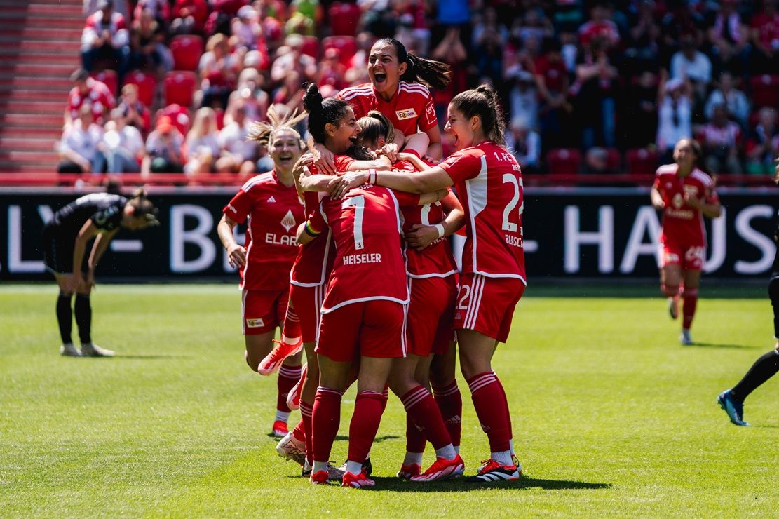 Spielerinnen eines Fußballteams in roten Trikots feiern einen Erfolg im Stadion, umgeben von jubelnden Fans.