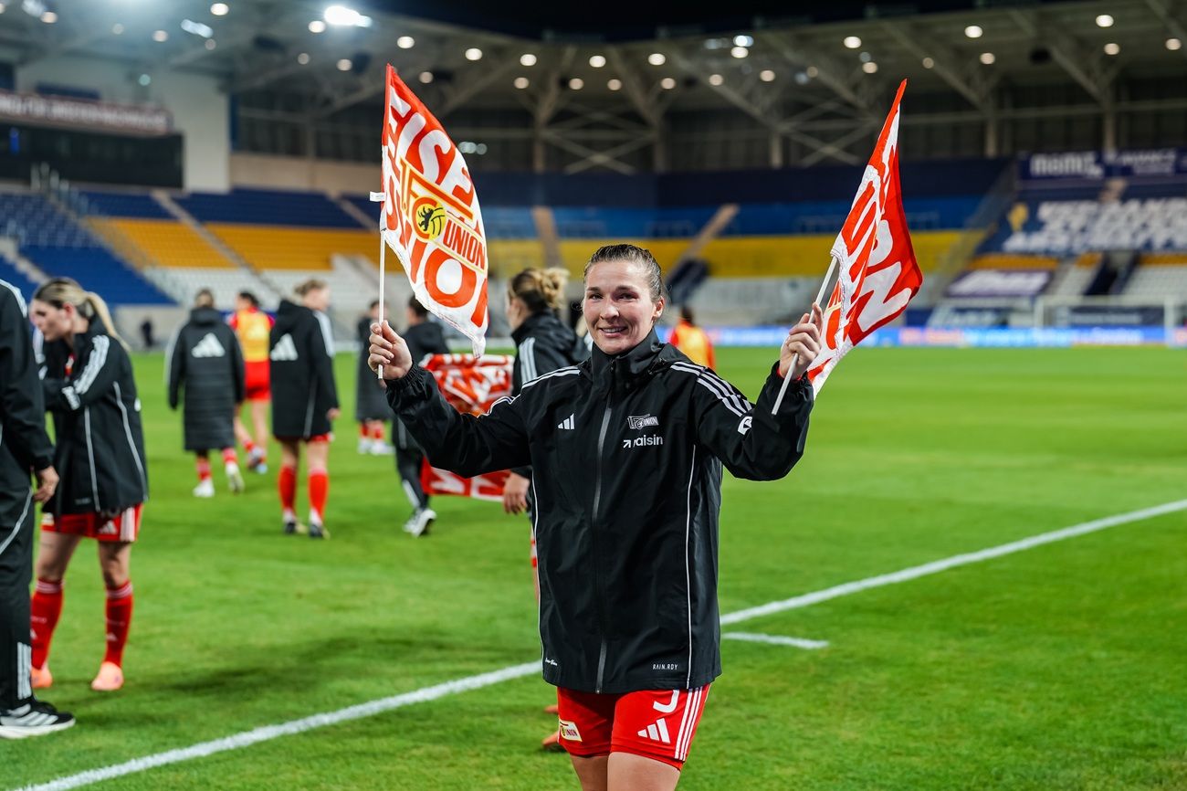 Eine sportliche Frau in schwarz-roter Kleidung hält zwei Fahnen und lächelt auf einem Fußballfeld nach einem Spiel.