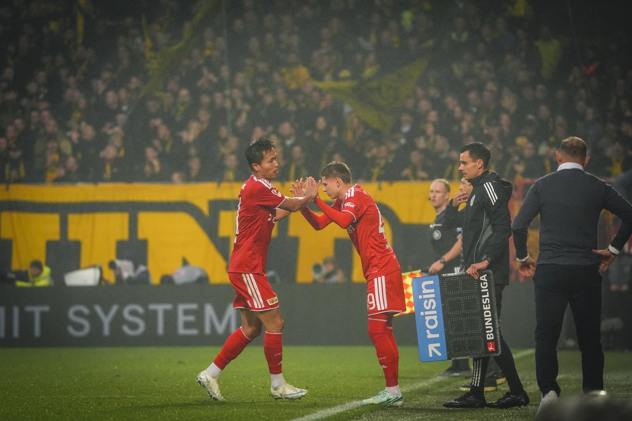 Zwei Fußballspieler in roten Trikots gratulieren sich während eines Spiels, im Hintergrund jubelnde Fans.