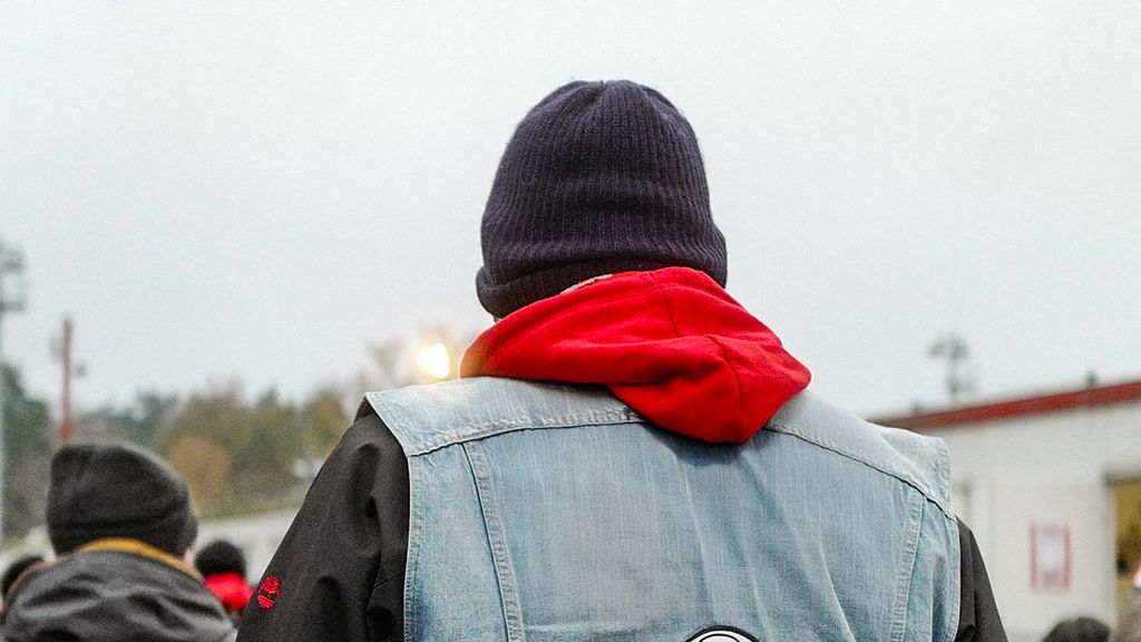 Person von hinten, trägt eine blaue Jeansjacke und eine rote Kapuzenjacke sowie eine schwarze Mütze, Szene bei grauem Wetter.