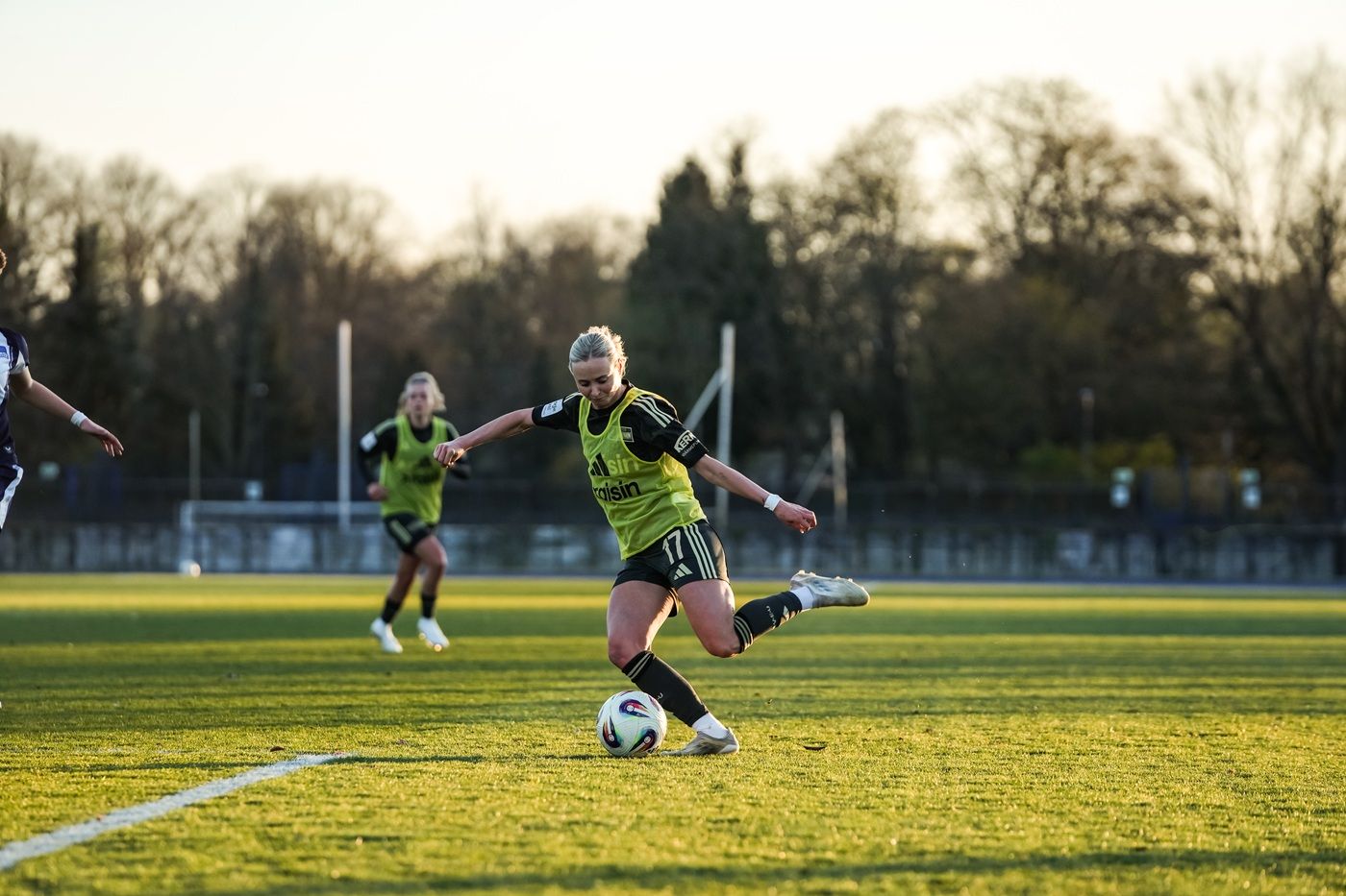 Eine Fußballspielerin in gelber Trainingsturniershirt schießt den Ball auf einem grünen Rasenplatz. Im Hintergrund eine weitere Spielerin.