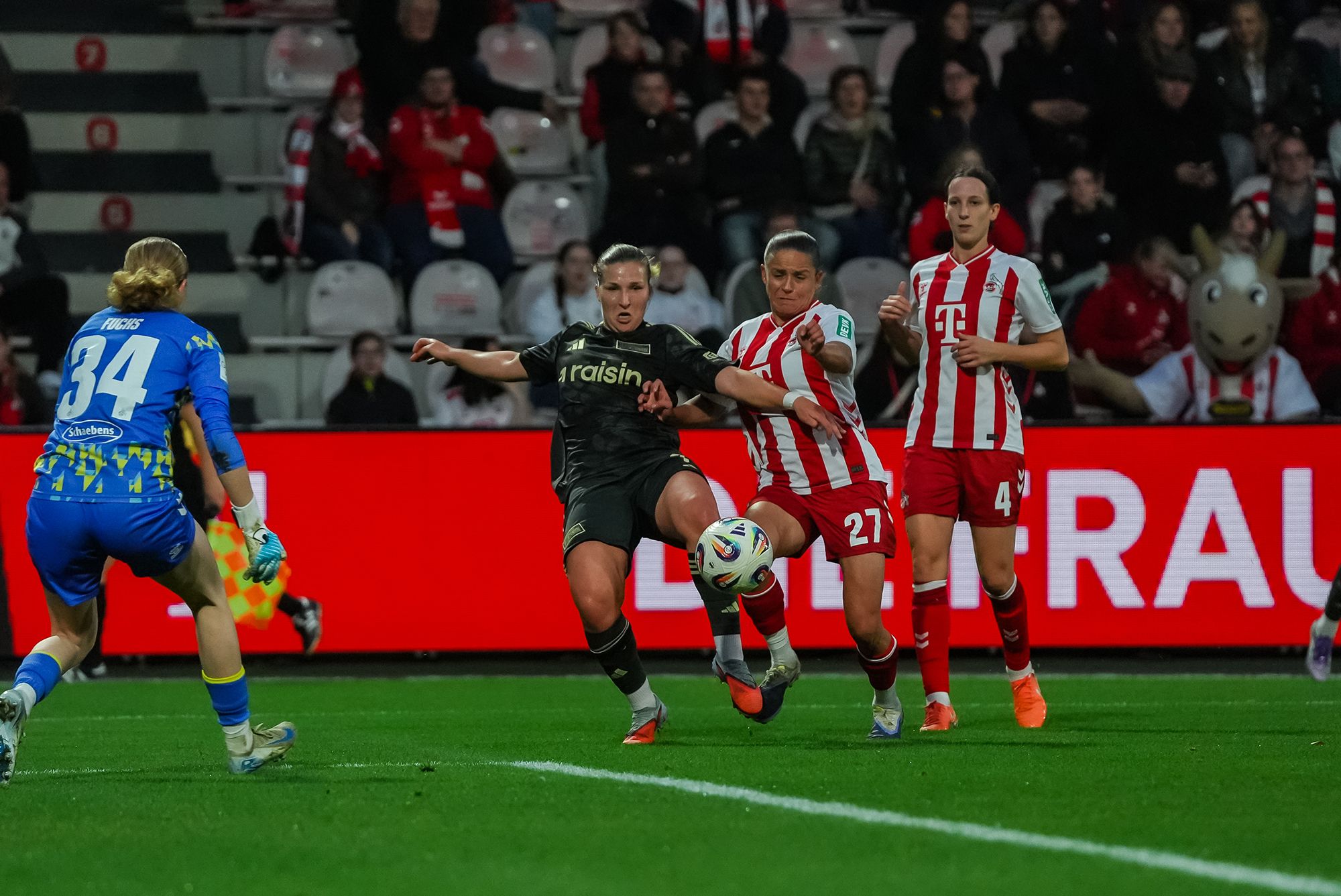 Spielerinnen kämpfen um den Ball im Fußballspiel, während ein Torwart in der Nähe positioniert ist. Fans sind im Hintergrund sichtbar.