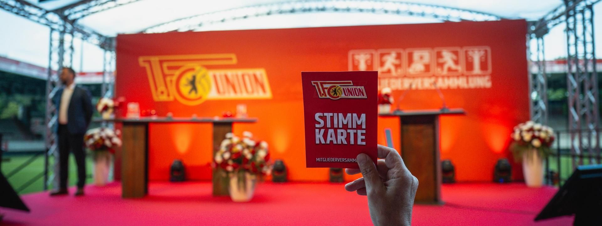 Hand hält eine rote Stimmkarte vor einer Bühne mit dekorativem Hintergrund und zwei Stehtischen in einem Stadion.