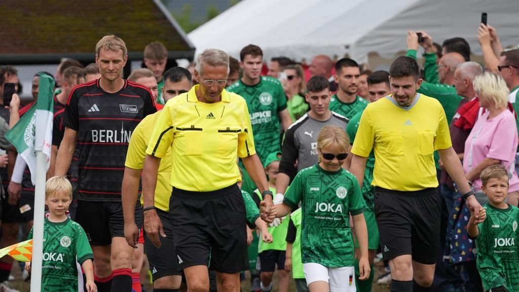 Eine Gruppe von Fußballspielern und Schiedsrichtern betritt das Spielfeld, begleitet von Kindern in Trikots.