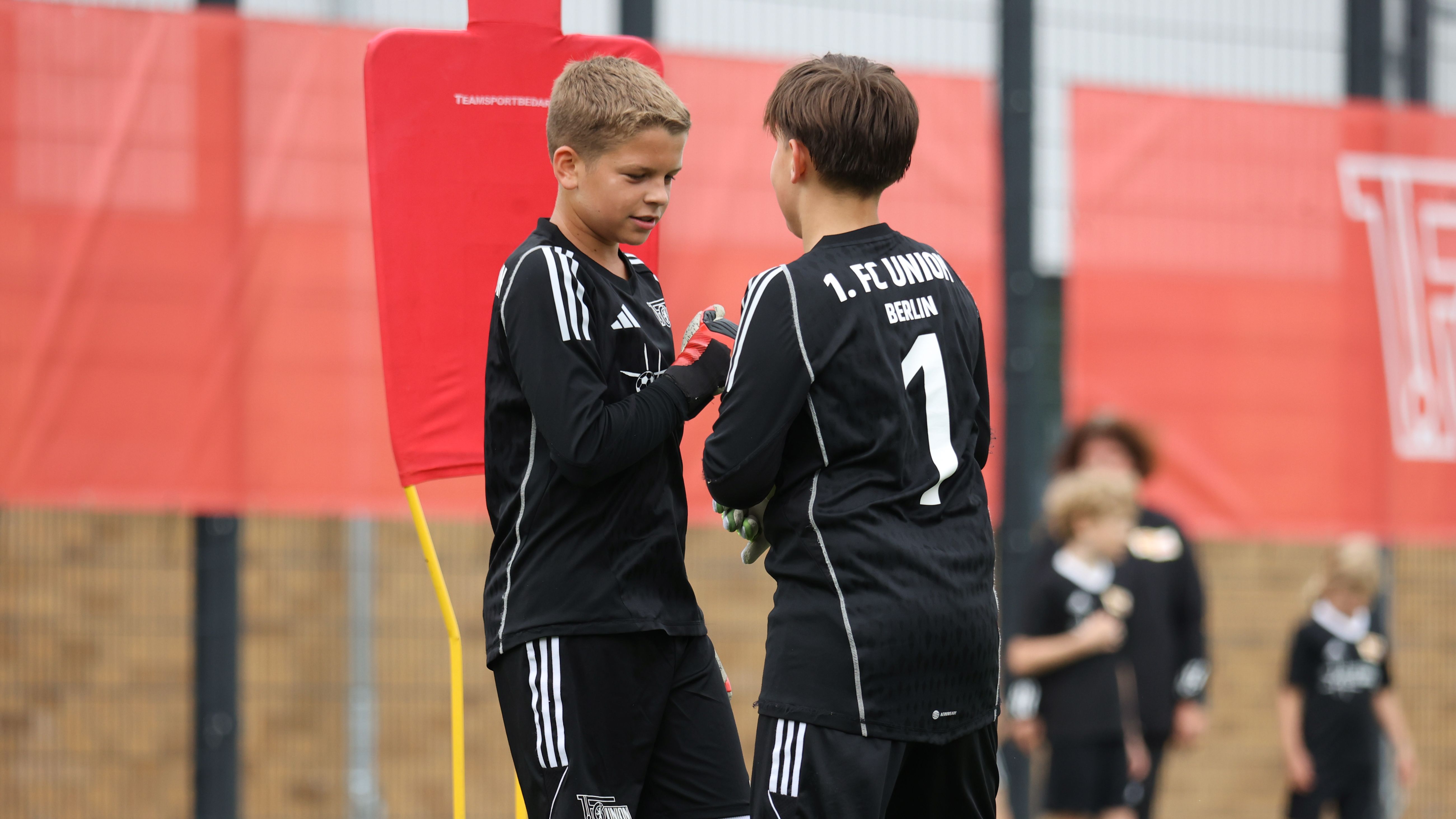 Zwei Jungen in schwarz-weißen Fußballtrikots stehen an einem Fußballfeld, einer trägt Handschuhe und spricht mit dem anderen.