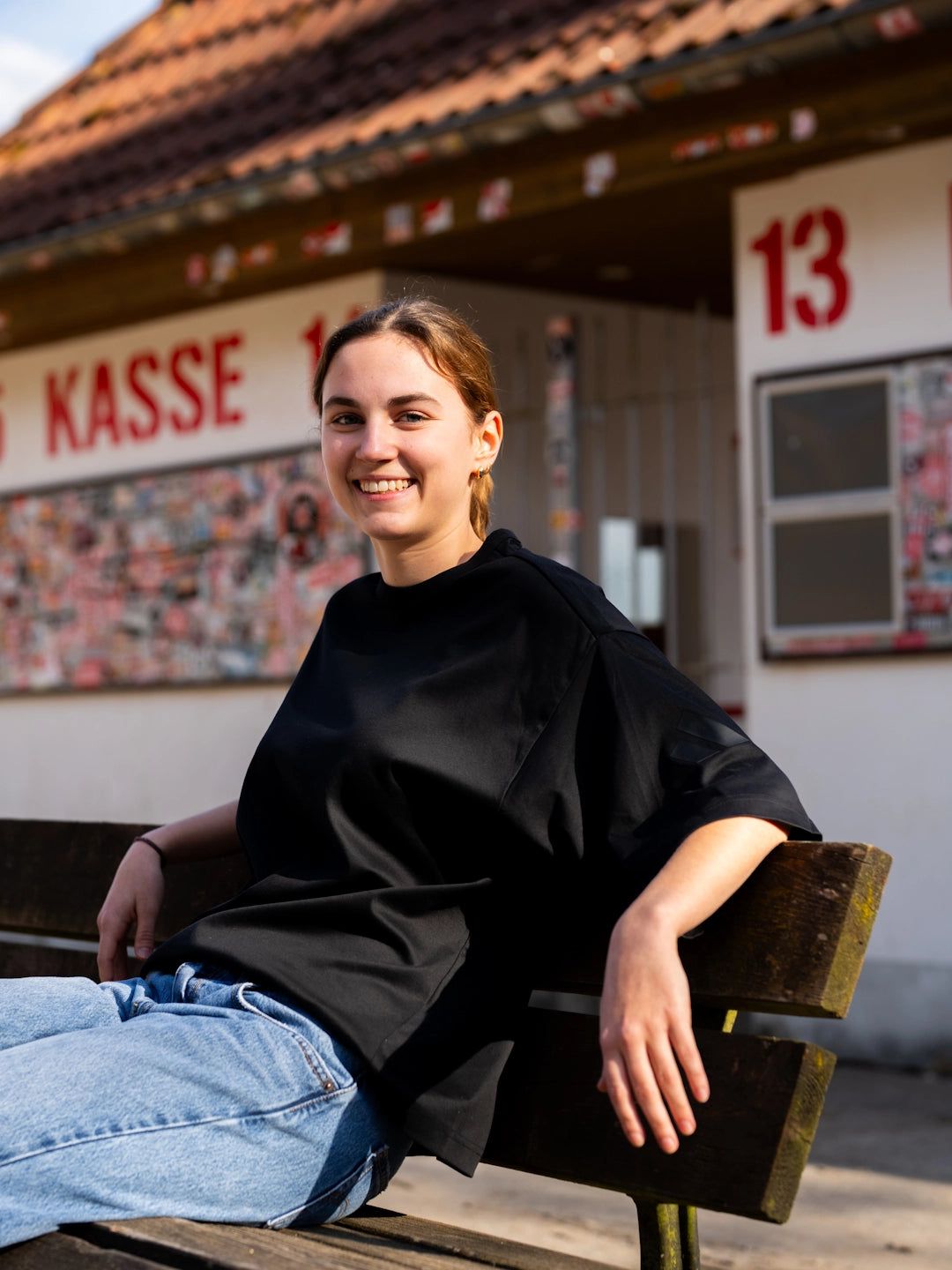 Junge Frau mit kurzer brauner Haarfrisur sitzt auf einer Bank und lächelt, im Hintergrund ein Gebäude mit der Aufschrift „KASSE“.