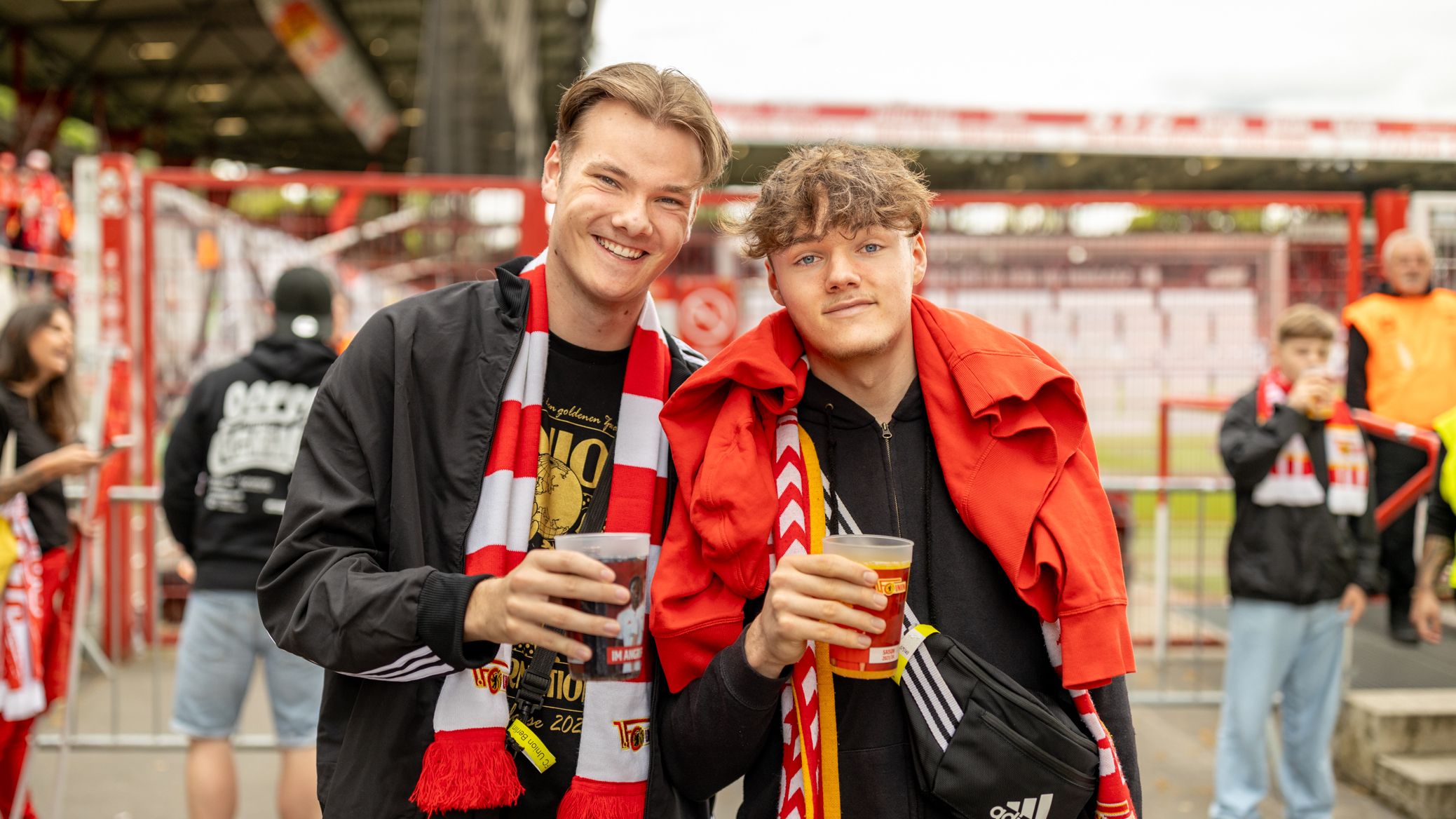 Zwei junge Männer stehen lächelnd am Stadioneingang, jeder hält ein Getränk in der Hand, umgeben von Fans in Fußballkleidung.