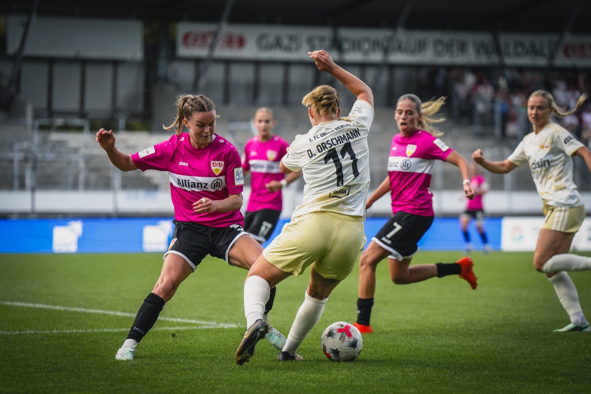 Zwei Fußballspielerinnen im intensiven Zweikampf auf dem Spielfeld, während andere Spielerinnen im Hintergrund aktiv sind.