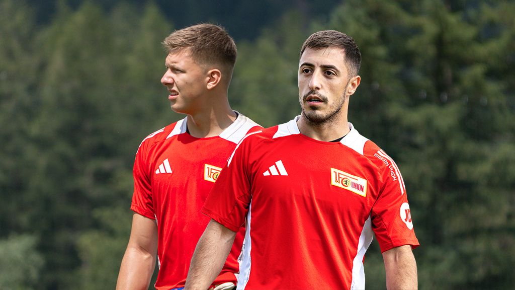 Zwei Fußballspieler in rot-weißen Trikots stehen auf einem Platz, umgeben von Bäumen im Hintergrund.