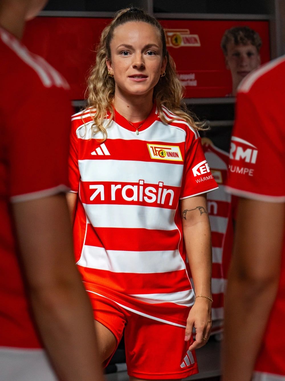 Eine Fußballspielerin trägt das Heimtrikot von Union Berlin und posiert in einer Umkleidekabine.