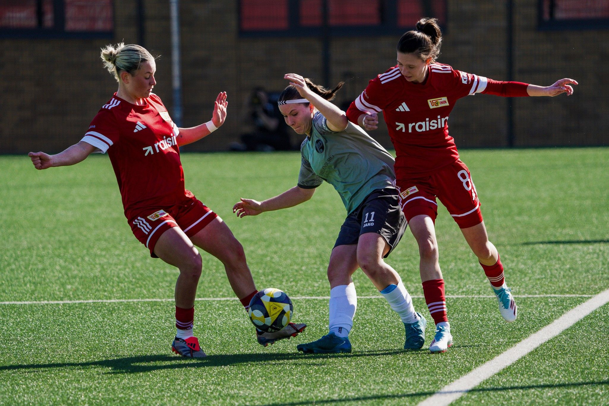 Zwei Spielerinnen im roten Trikot verteidigen gegen eine Spielerin im grauen Trikot, während der Ball im Spiel ist.