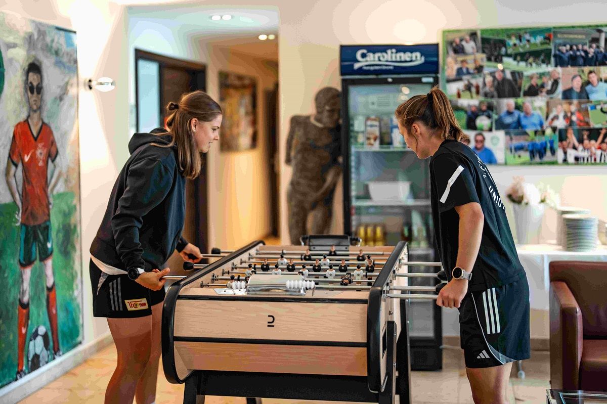 Zwei Frauen spielen Tischfußball in einem Raum mit Fotos an der Wand und einer Statue im Hintergrund.