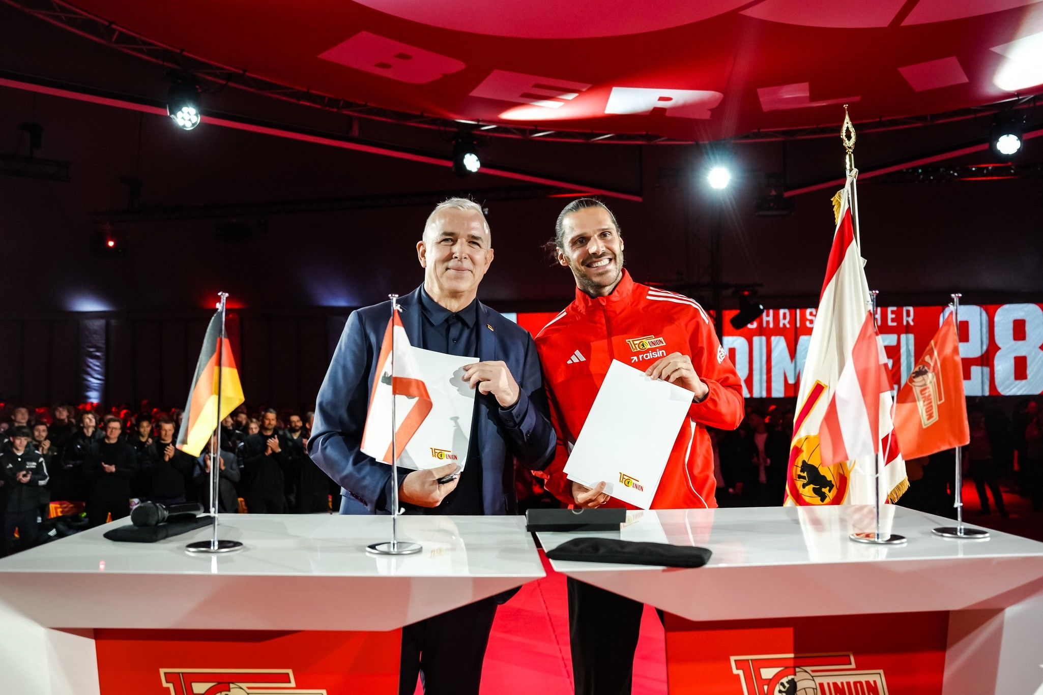 Zwei Personen in Anzügen unterschreiben Verträge an einem Tisch mit Flaggen im Hintergrund, während eine Menschenmenge zuschaut.