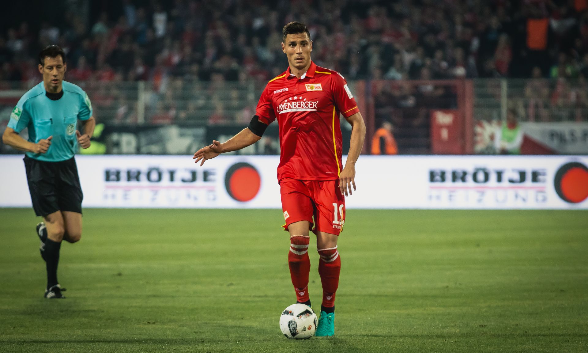 Ein Fußballspieler in roter Trikot steht auf dem Spielfeld, während ein Schiedsrichter im Hintergrund beobachtet. Fans sind im Stadion sichtbar.