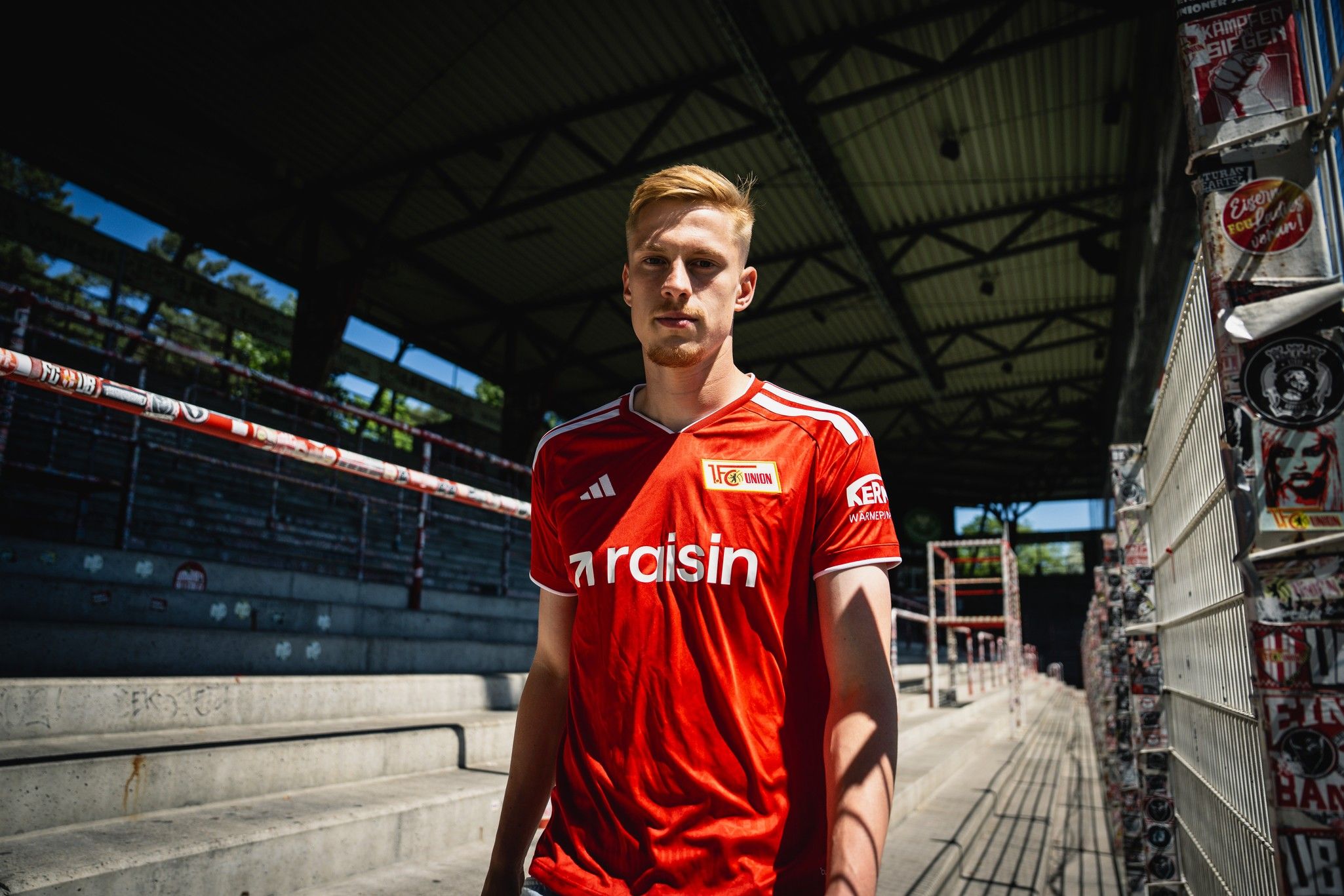Ein junger Mann in einem roten Fußballtrikot steht in einem Stadion mit leeren Sitzen im Hintergrund.