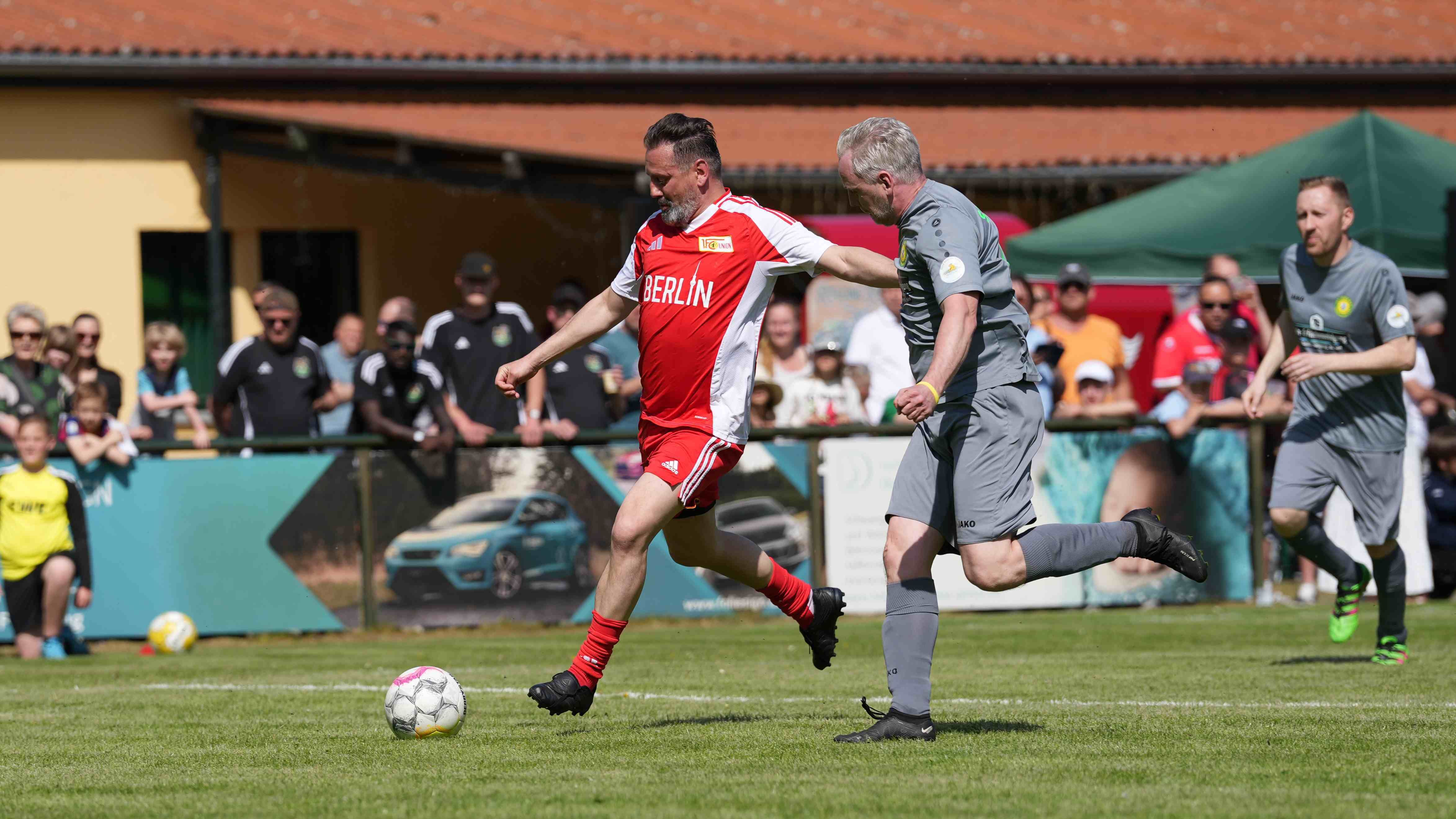 Ein Fußballspieler in rotem Trikot dribbelt den Ball, während ein Gegner in grauem Trikot ihm folgt. Im Hintergrund sind Zuschauer zu sehen.