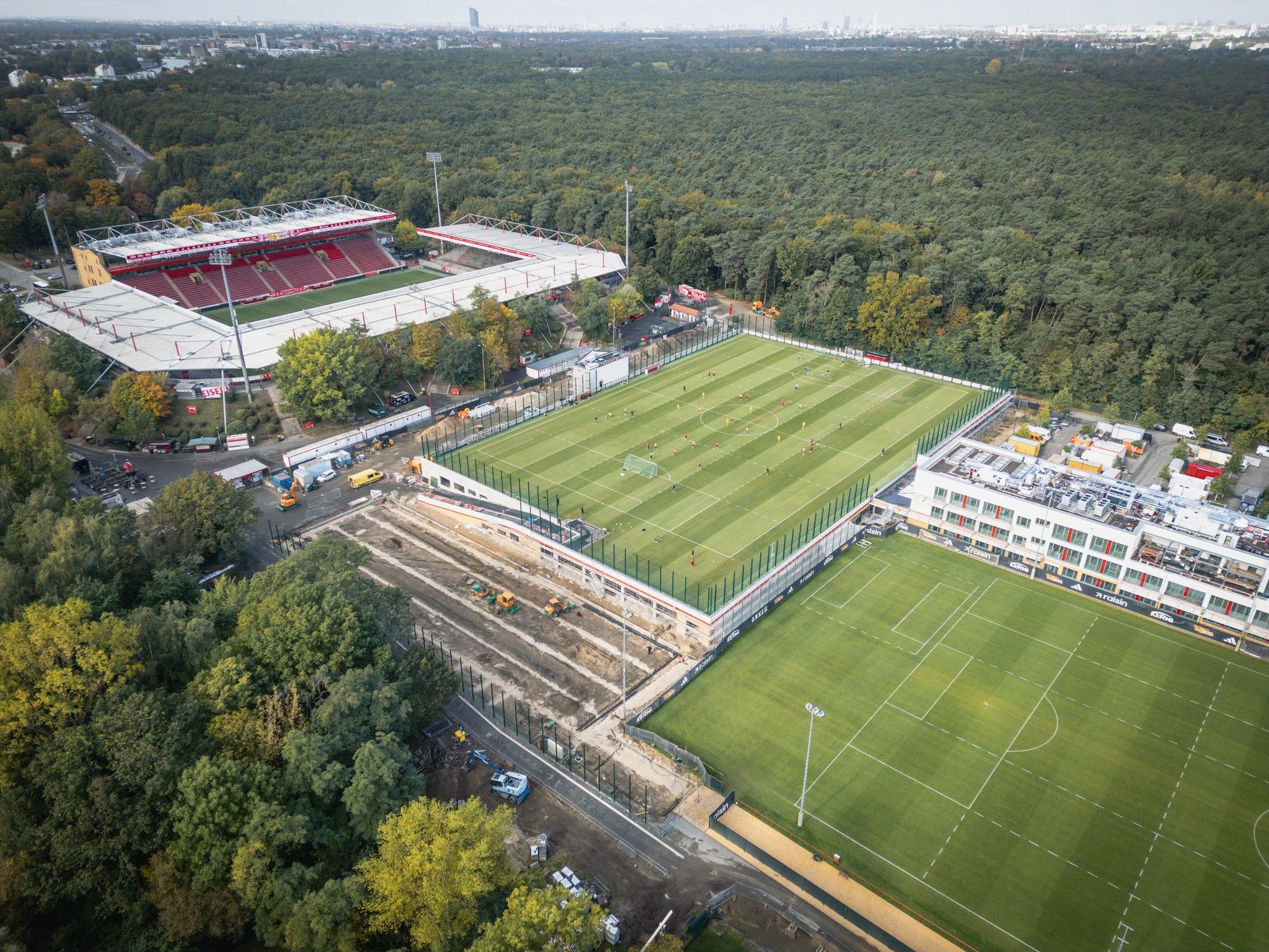 Luftaufnahme eines Sportgeländes mit mehreren Fußballfeldern, umgeben von Bäumen und einem Stadion im Hintergrund.