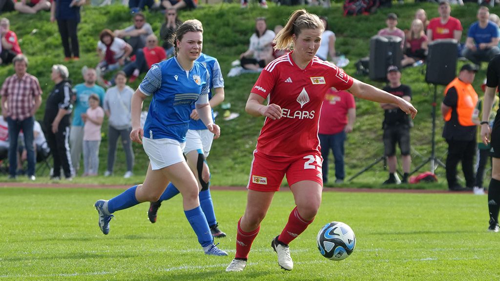 A female soccer player in a red jersey dribbles the ball on a green field, while a player in a blue jersey follows her.