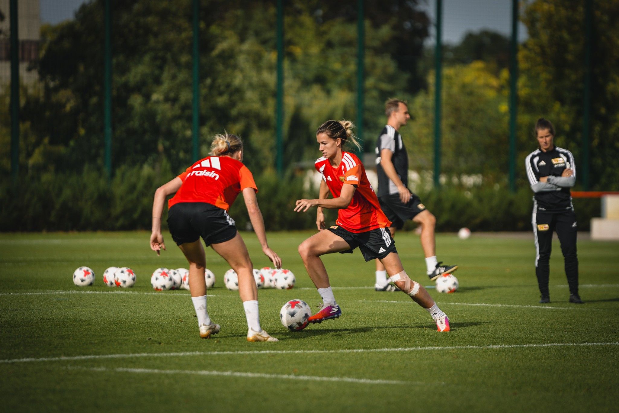 Zwei Fußballspielerinnen in roten Trikots üben auf einem Trainingsplatz, während andere Spieler im Hintergrund trainieren.