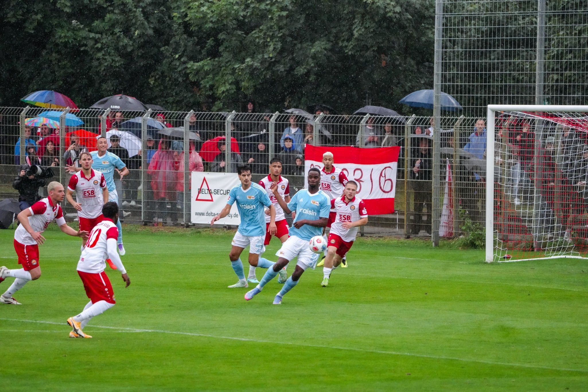 Spielszene eines Fußballspiels im Regen, Spieler in roten und blauen Trikots, Zuschauer mit Regenschirmen im Hintergrund.