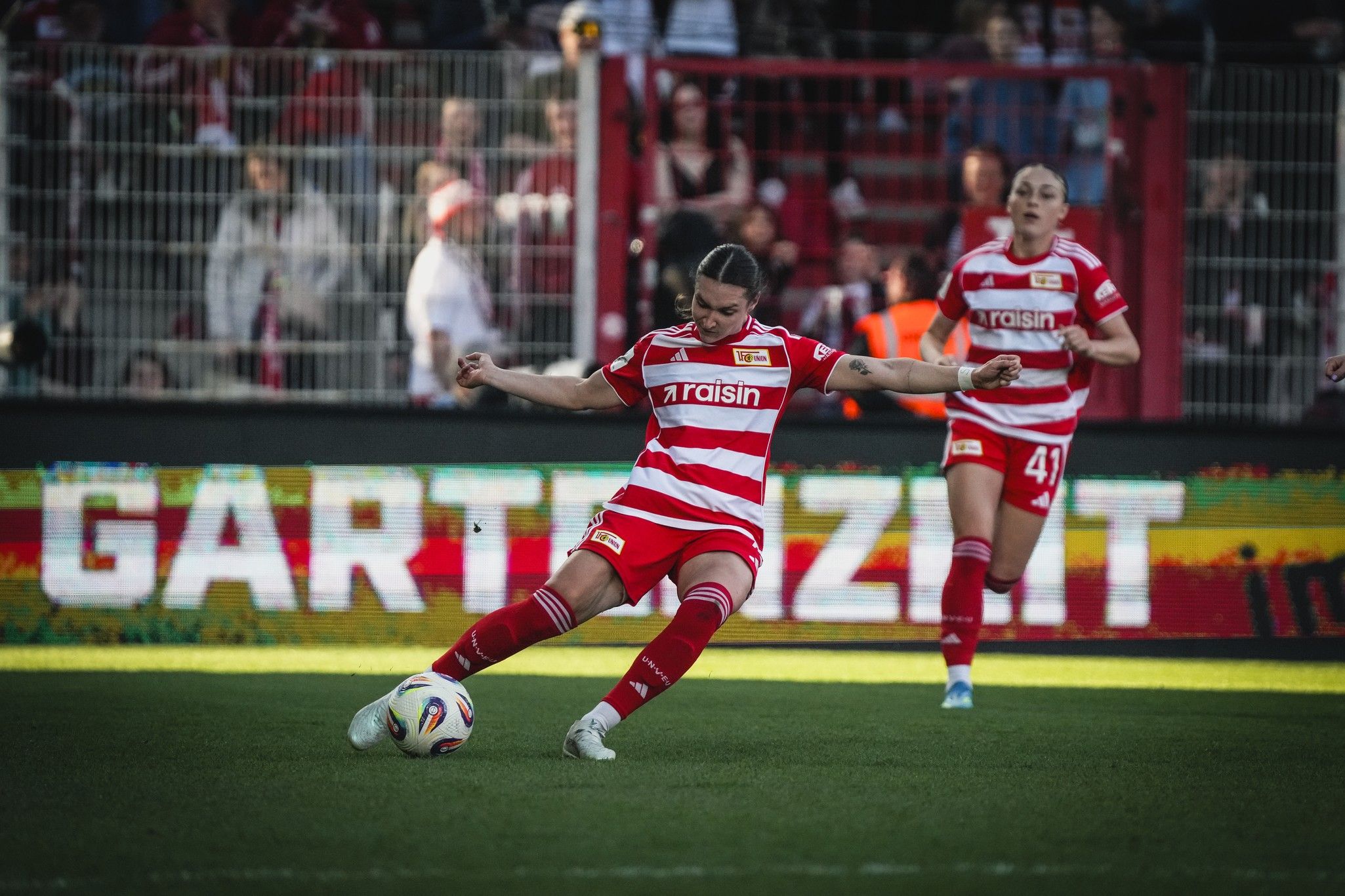Spielerin in rotem und weißem Trikot führt einen Schuss aus, während zwei Teamkolleginnen im Hintergrund stehen.